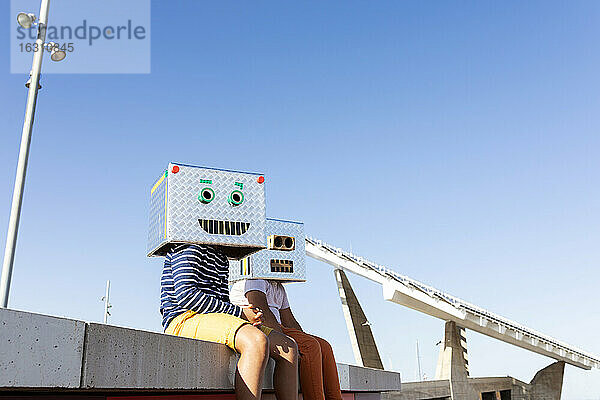 Jungen mit Robotermasken aus Kartons  die auf einer Stützmauer vor blauem Himmel sitzen