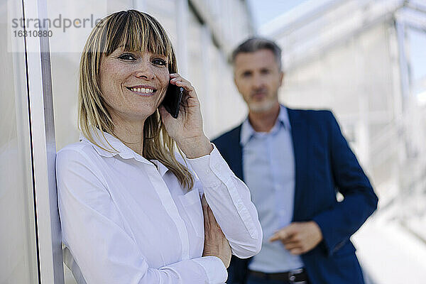 Lächelnde Geschäftsfrau im Gespräch über ein Smartphone  während ein männlicher Mitarbeiter im Hintergrund steht