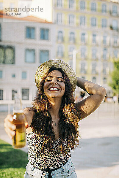 Fröhliche junge Frau mit Hut  die eine Bierflasche hält  während sie in der Stadt steht