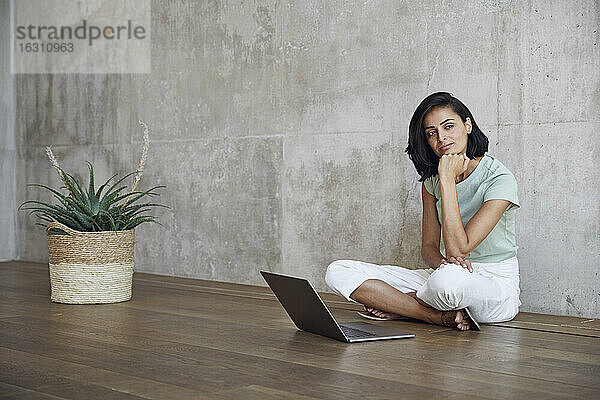 Nachdenkliche Unternehmerin sitzt mit Laptop auf Hartholzboden an der Wand im Büro