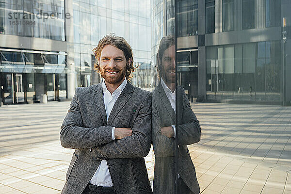 Gut aussehender Geschäftsmann  der mit verschränkten Armen vor dem Spiegelbild eines Bürogebäudes im Finanzviertel steht
