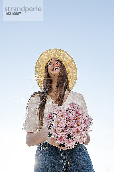 Lachende Frau steht und hält rosa Blumen gegen den klaren Himmel