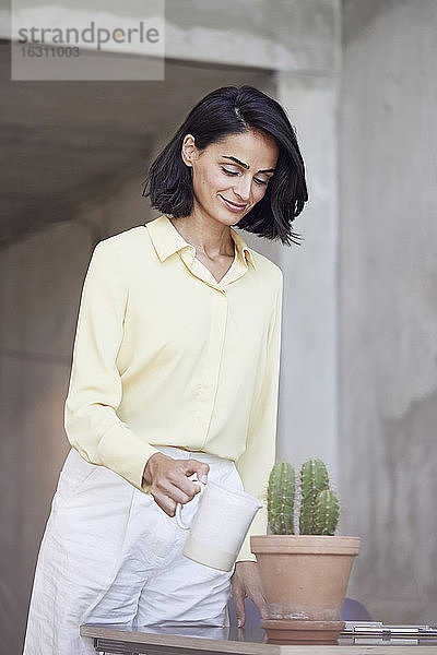 Lächelnde Geschäftsfrau  die eine Kaktuspflanze auf dem Schreibtisch im Büro gießt
