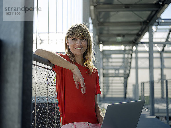 Lächelnde Geschäftsfrau  die einen Laptop benutzt  während sie an einem Geländer im Gewächshaus sitzt