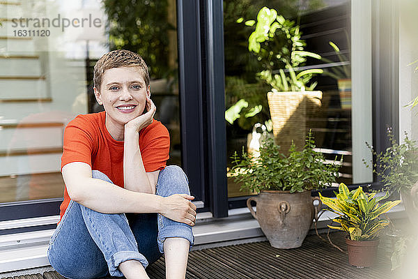 Lächelnde schöne Frau mit Hand auf dem Kinn sitzt gegen Haus im Hof