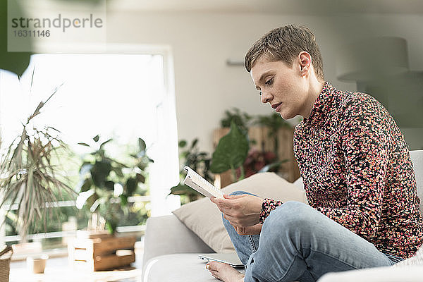 Mittlere erwachsene Frau mit kurzen Haaren liest ein Buch  während sie zu Hause auf dem Sofa sitzt