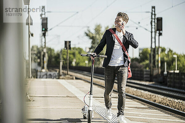 Lächelnder Mann  der mit einem Mobiltelefon spricht  während er mit einem Roller auf dem Bahnsteig spazieren geht
