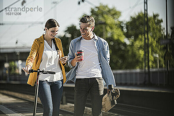 Junge Frau zeigt einem männlichen Freund beim Spaziergang am Bahnhof ihr Smartphone