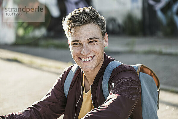 Lächelnder junger Mann in Freizeitkleidung auf der Straße sitzend