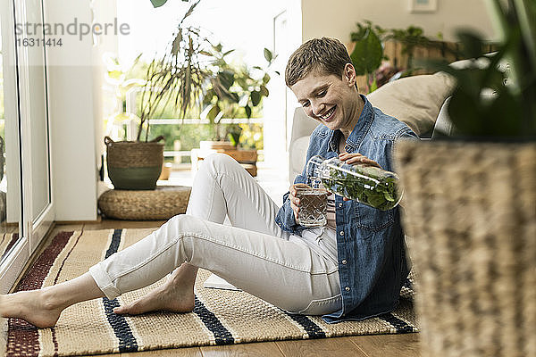 Lächelnde Frau mit kurzen Haaren gießt ein Getränk in ein Glas  während sie zu Hause auf einem Teppich sitzt