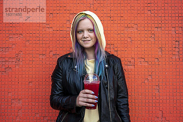 Porträt einer jungen Frau mit gefärbtem Haar und einem Getränk zum Mitnehmen vor einer roten Wand