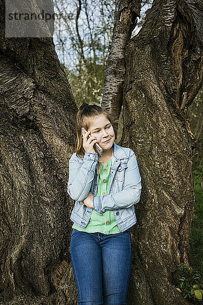 Lächelndes Mädchen  das mit seinem Smartphone spricht  während es gegen einen Baumstamm im Park steht
