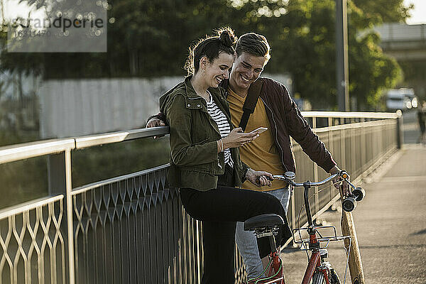 Lächelndes junges Paar  das auf sein Smartphone schaut  während es mit dem Fahrrad in der Stadt steht