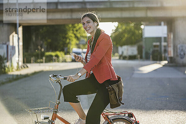 Glückliche junge Frau  die ihr Smartphone benutzt  während sie auf einer Straße in der Stadt Fahrrad fährt