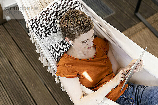 Mittlere erwachsene Frau mit kurzen Haaren  die ein digitales Tablet benutzt  während sie auf einer Hängematte auf der Veranda liegt