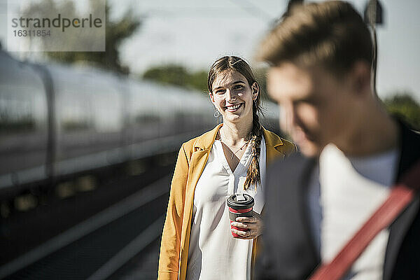 Lächelnde junge Frau mit wiederverwendbarem Kaffeebecher am Bahnhof
