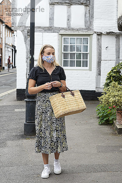 Junge blonde Frau mit Gesichtsmaske geht mit einer Einkaufstasche durch das Dorf.
