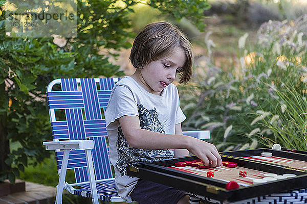 Ein kleiner Junge spielt Backgammon im Freien in einem Garten.