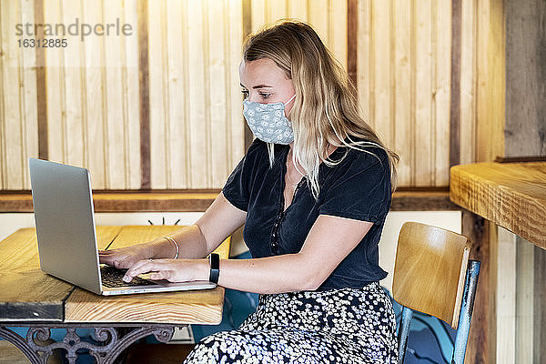 Junge blonde Frau mit blauer Gesichtsmaske  die am Tisch sitzt und einen Laptop-Computer benutzt.