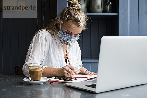 Frau mit Gesichtsmaske sitzt allein an einem Café-Tisch mit einem Laptop  der ferngesteuert arbeitet.