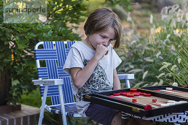 Ein kleiner Junge spielt Backgammon im Freien in einem Garten.