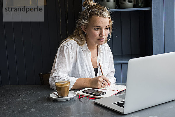 Frau  die allein an einem Café-Tisch mit einem Laptop-Computer sitzt  in ein Notizbuch schreibt und aus der Ferne arbeitet.