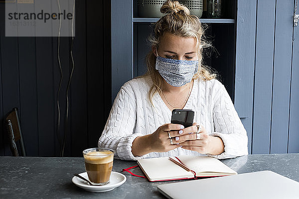 Frau mit Gesichtsmaske sitzt allein an einem Café-Tisch mit einem Laptop-Computer und benutzt ein Mobiltelefon
