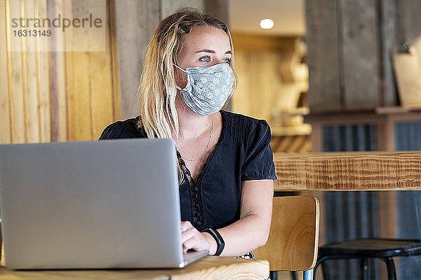 Junge blonde Frau mit blauer Gesichtsmaske  die am Tisch sitzt und einen Laptop-Computer benutzt.