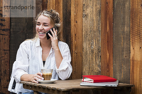 Junge blonde Frau  die allein in einem Café sitzt  ein Mobiltelefon benutzt und aus der Ferne arbeitet.