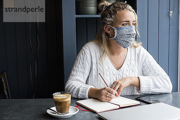 Frau mit Gesichtsmaske  die allein an einem Café-Tisch sitzt und einen Laptop benutzt  der aus der Ferne arbeitet.