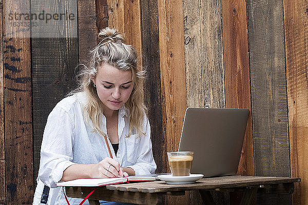 Junge blonde Frau  die allein an einem Café-Tisch mit einem Laptop sitzt und aus der Ferne arbeitet.