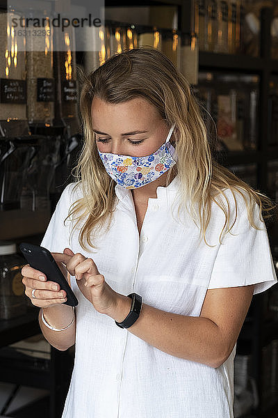 Frau trägt Gesichtsmaske  kauft in einem abfallfreien örtlichen Geschäft ein und benutzt ein Telefon