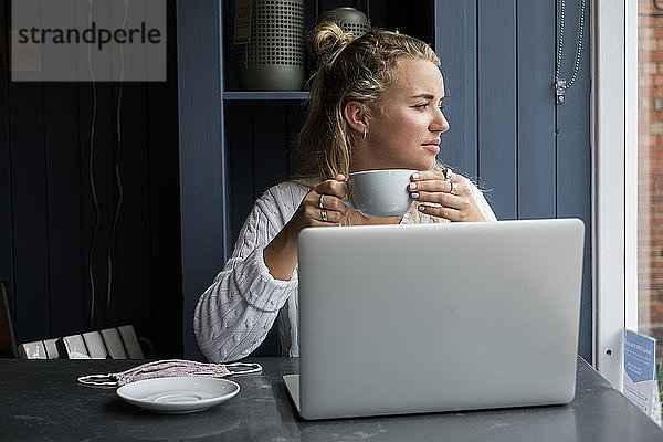 Junge blonde Frau  die allein an einem Cafétisch mit einem Laptop-Computer sitzt und aus der Ferne arbeitet.