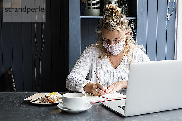 Frau mit Gesichtsmaske sitzt allein mit einem Laptop  schreibt in ein Notizbuch  arbeitet aus der Ferne.