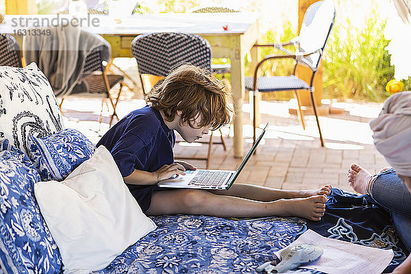 Junge mit braunen Haaren sitzt auf einem Bett im Freien und macht Hausaufgaben am Laptop.