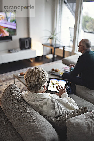 Hochwinkelansicht einer älteren Frau mit digitalem Tablet  während der männliche Partner auf dem Sofa im Wohnzimmer sitzt