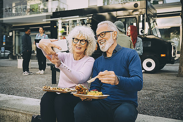 Lächelnde ältere Frau  die sich mit einem Mann beim Essen gegen einen Speisewagen in der Stadt vergnügt