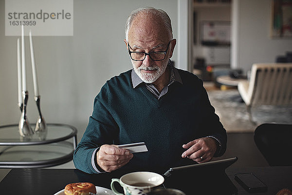 Runzliger Mann hält Kreditkarte per digitalem Tablet  während er am Esstisch im Wohnzimmer sitzt
