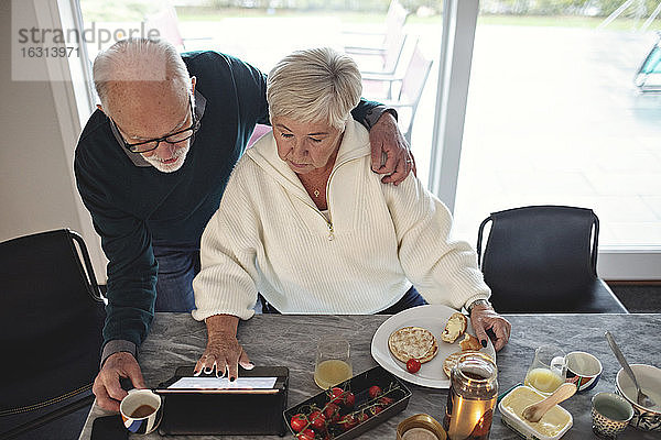 Hochwinkelansicht eines älteren Paares  das auf ein digitales Tablet schaut  während es am Esstisch im Wohnzimmer sitzt