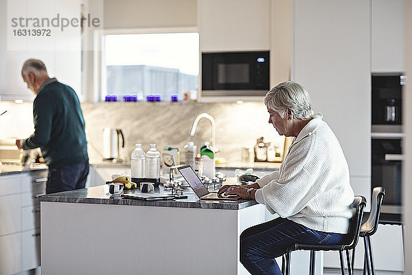 Ältere Frau benutzt Laptop  während ein Mann in der Küche Essen zubereitet
