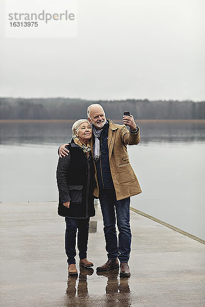 Älteres Ehepaar in voller Länge beim Selfie am Seeufer vor klarem Himmel