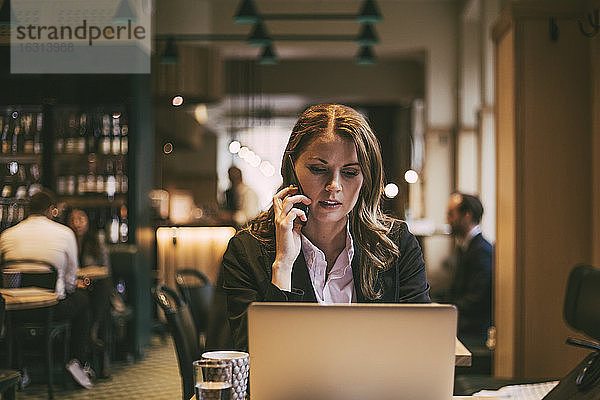 Geschäftsfrau telefoniert  während sie im Restaurant auf den Laptop schaut