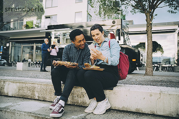 Lächelnde Freunde  die sich ein Smartphone teilen  während sie beim Essen gegen einen Speisewagen in der Stadt sitzen