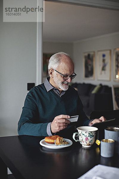 Lächelnder  faltiger Mann hält Kreditkarte  während er im Wohnzimmer ein digitales Tablett über dem Tisch benutzt