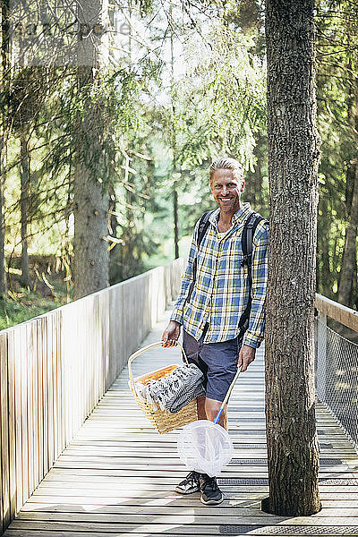 Porträt eines lächelnden Mannes mit Picknickkorb auf einem Steg im Wald stehend