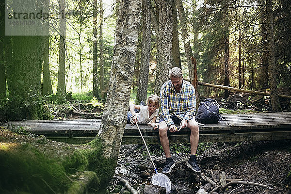 Vater mit Rucksack unterhält sich mit der Tochter  während er auf einem Steg im Wald sitzt