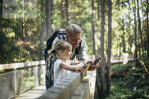 Lächelnder Vater mit Rucksack und Tochter beim Blick auf Smartphone im Wald