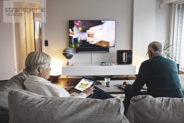 Ältere Frau benutzt digitales Tablet  während ihr Partner auf dem Sofa im Wohnzimmer sitzt