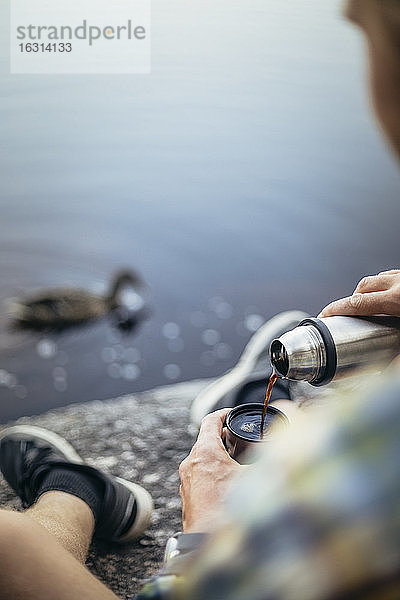 Reifer Mann gießt Kaffee ein  während er am See sitzt
