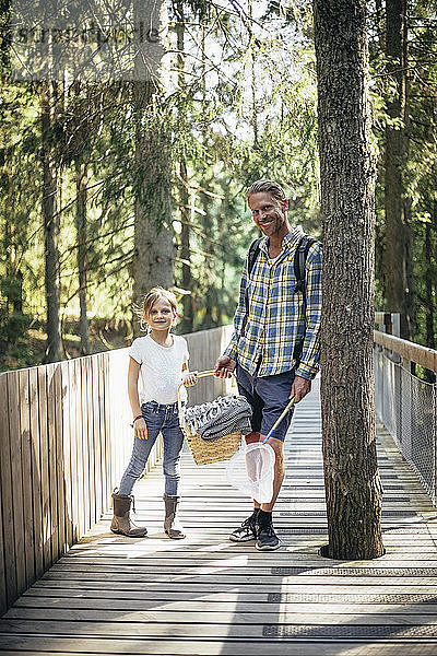 Porträt von Vater und Tochter mit Picknickkorb auf einem Steg im Wald stehend